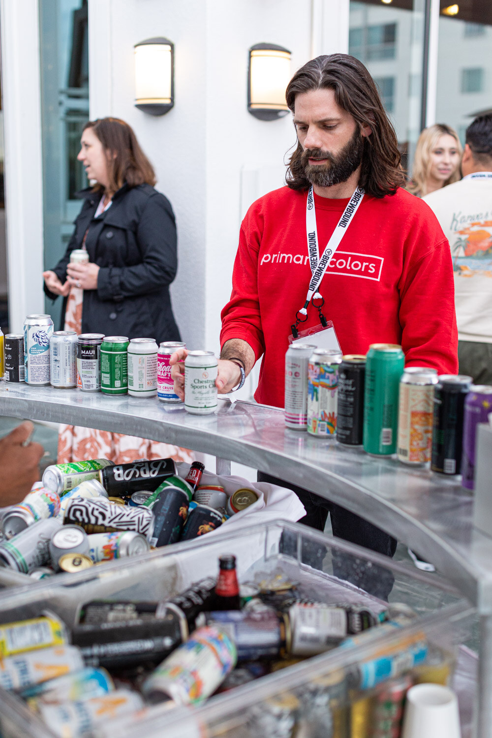 A man with long hair in a red sweatshirt reading primary colors stands behind a round counter of canned beverages and bins.