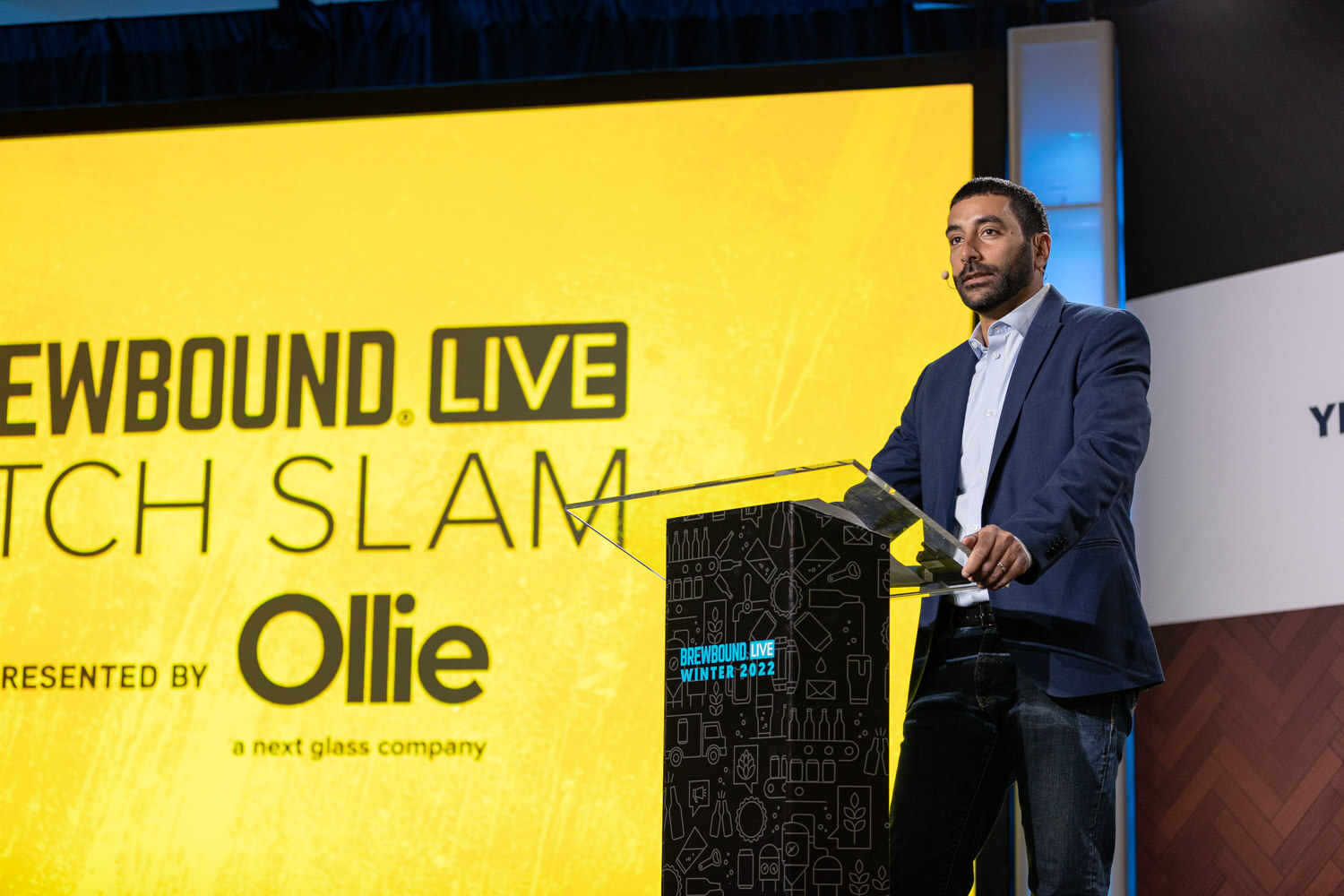 A man in a navy blazer stands at a podium in front of a yellow screen reading Brewbound Live Pitch Slam presented by Ollie.
