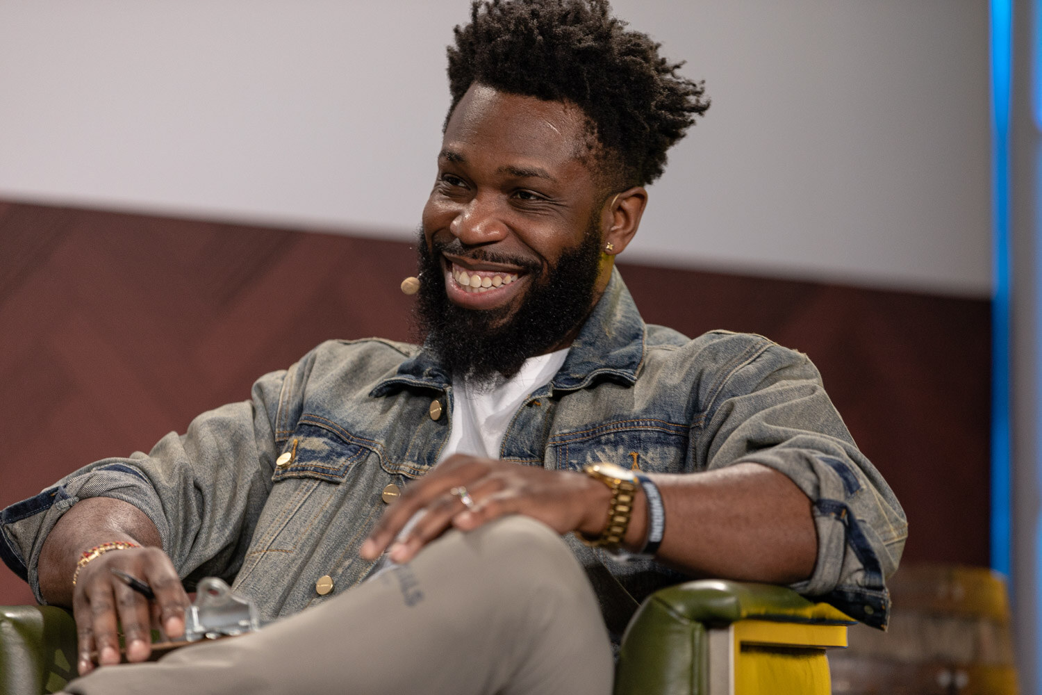 A smiling bearded man in a denim jacket sits back in a green armchair with his hand on his knee.