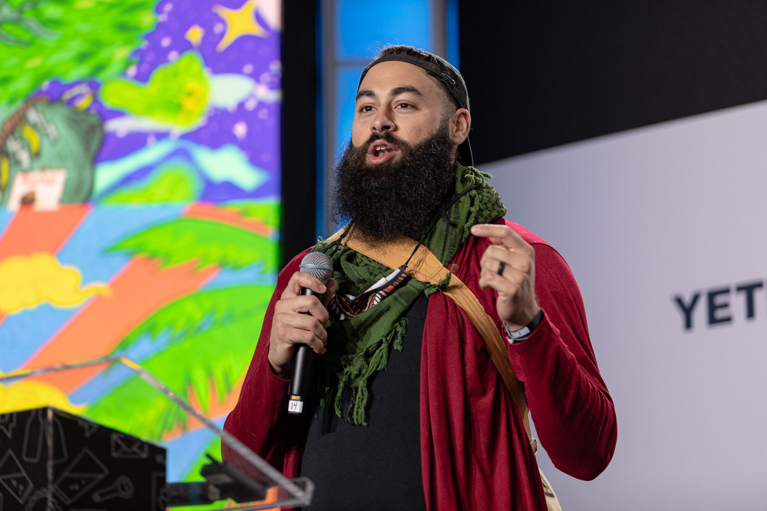 A bearded man in a red cardigan and green scarf holds a microphone in front of a colorful painted landscape on the screen.