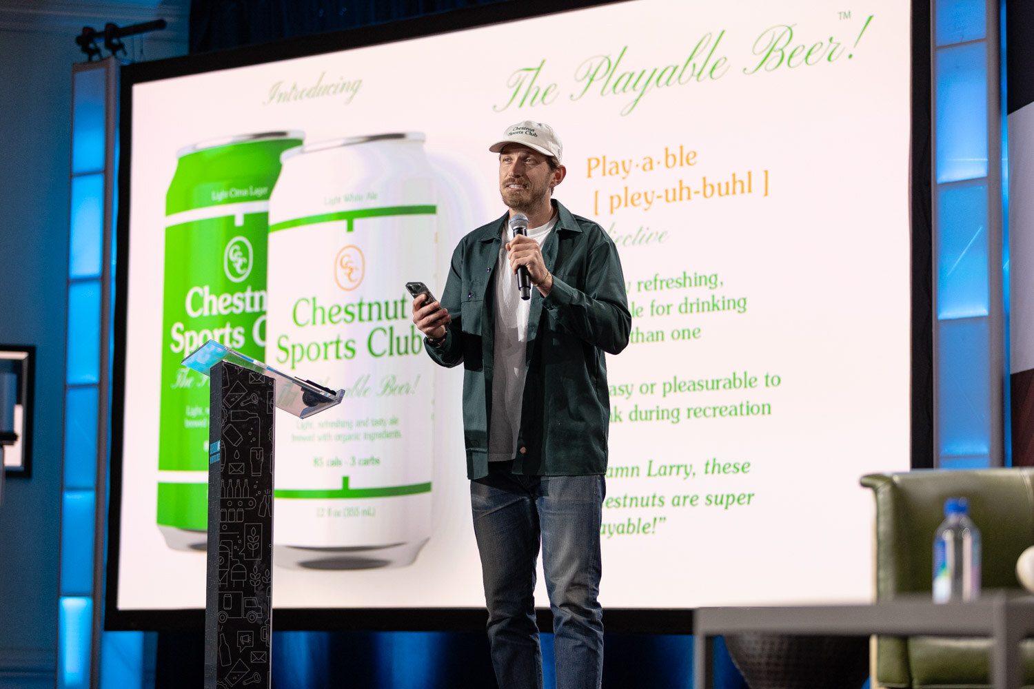 A man in a white cap and green jacket holds a microphone and phone beside a large screen showing green and white Chestnut Sports Club cans.