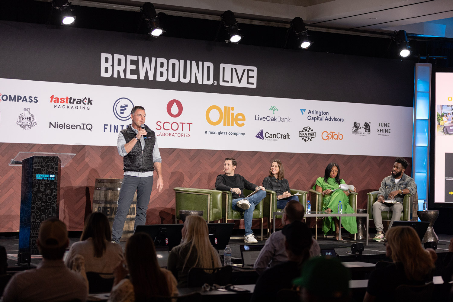Wide view of the Brewbound Live stage with a man at the podium and four judges seated in green armchairs.