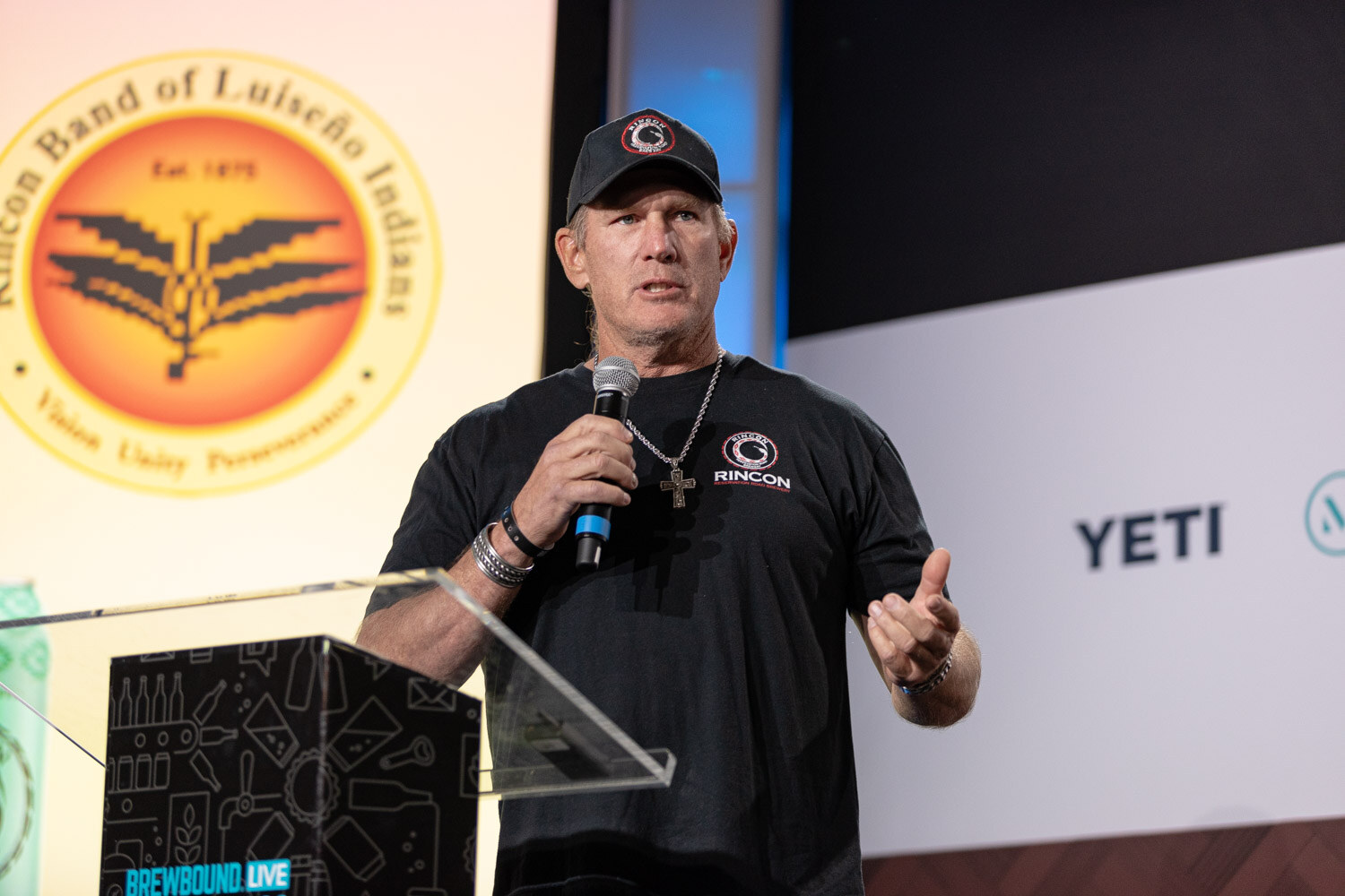 A man in a black cap and black T-shirt speaks into a microphone in front of a large Rincon Band of Luiseno Indians seal.