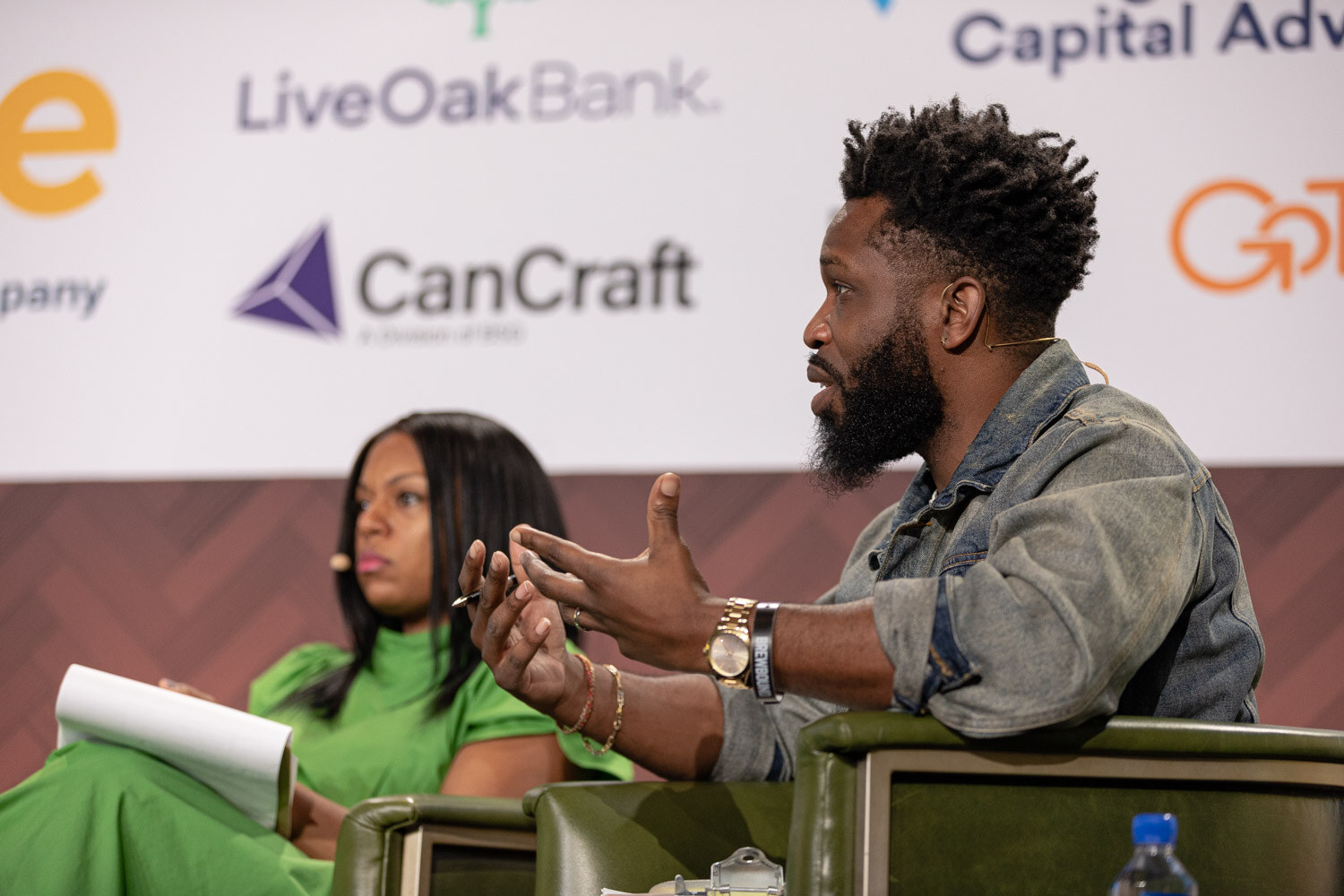 A bearded man in a denim jacket speaks with open hands next to a woman in a bright green dress, both in green armchairs.