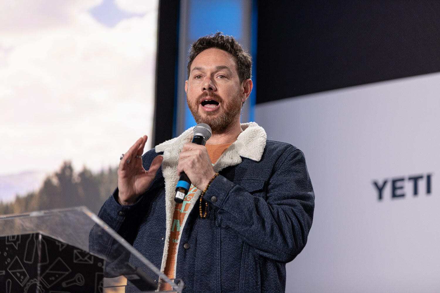 A man with dark hair in a denim jacket with a fleece collar holds a microphone behind a clear podium with a desert landscape on the screen behind him.