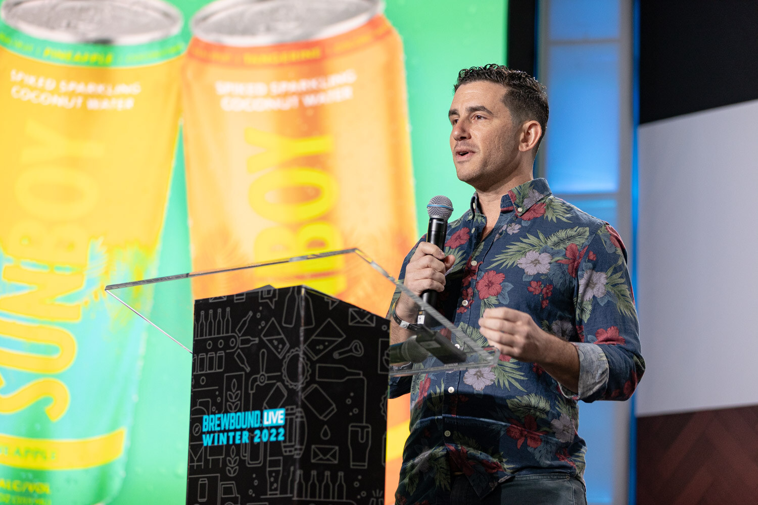 A man in a navy floral shirt holds a microphone at a podium with a green and orange slide showing Sunboy cans.