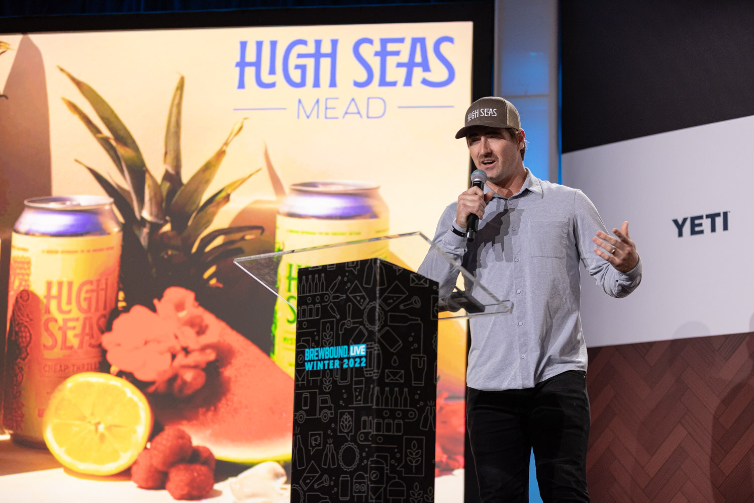 A man in a gray shirt and cap holds a microphone at a podium next to a slide reading High Seas Mead with pineapple and raspberries.