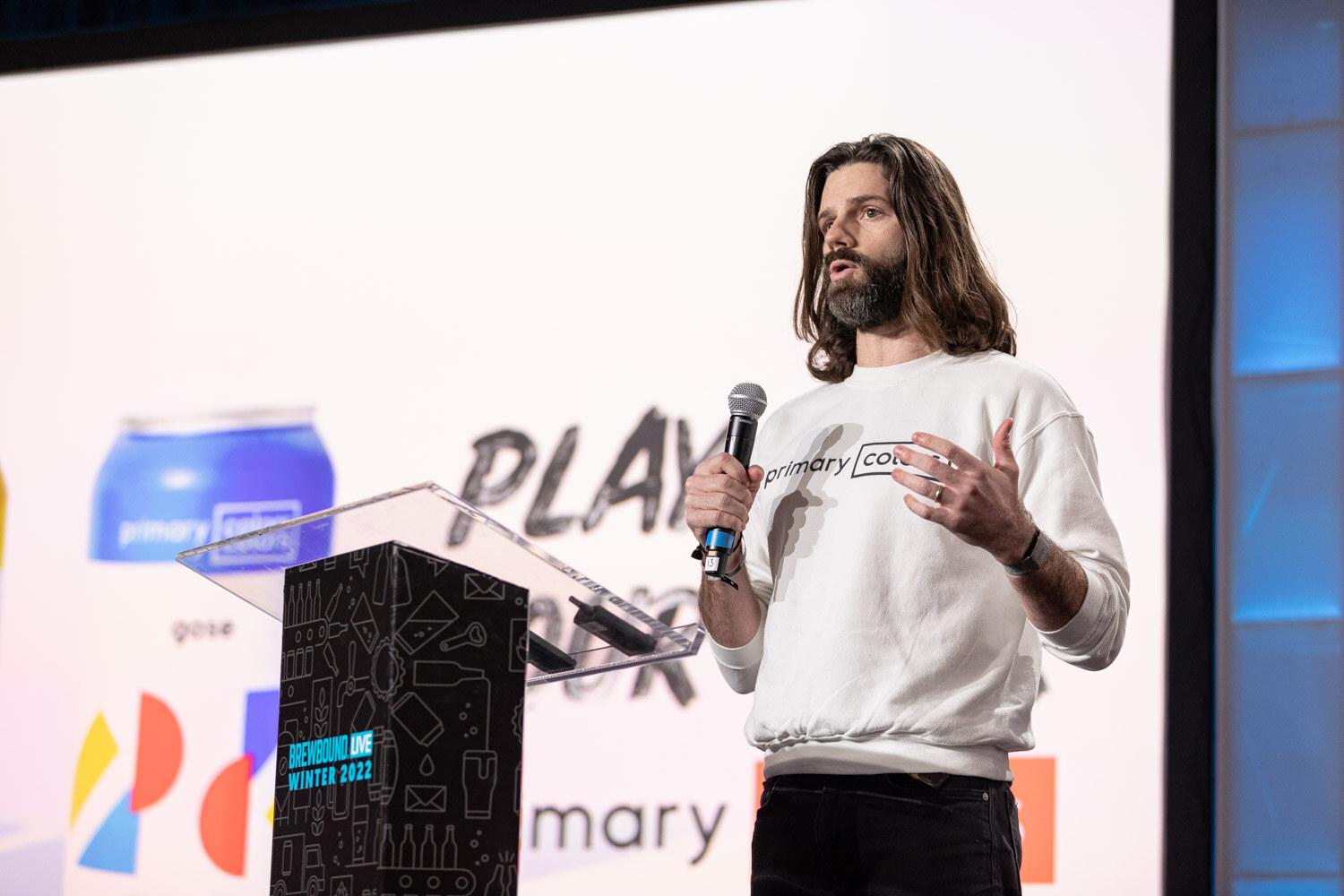 A man with long hair and a beard in a white sweatshirt reading primary colors holds a microphone beside a podium and a slide with a blue can.