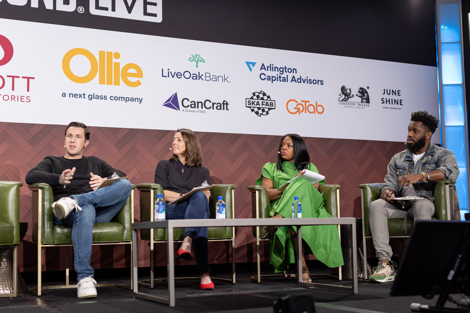 Wide view of four people in green armchairs on a stage under the Brewbound Live sponsor wall.
