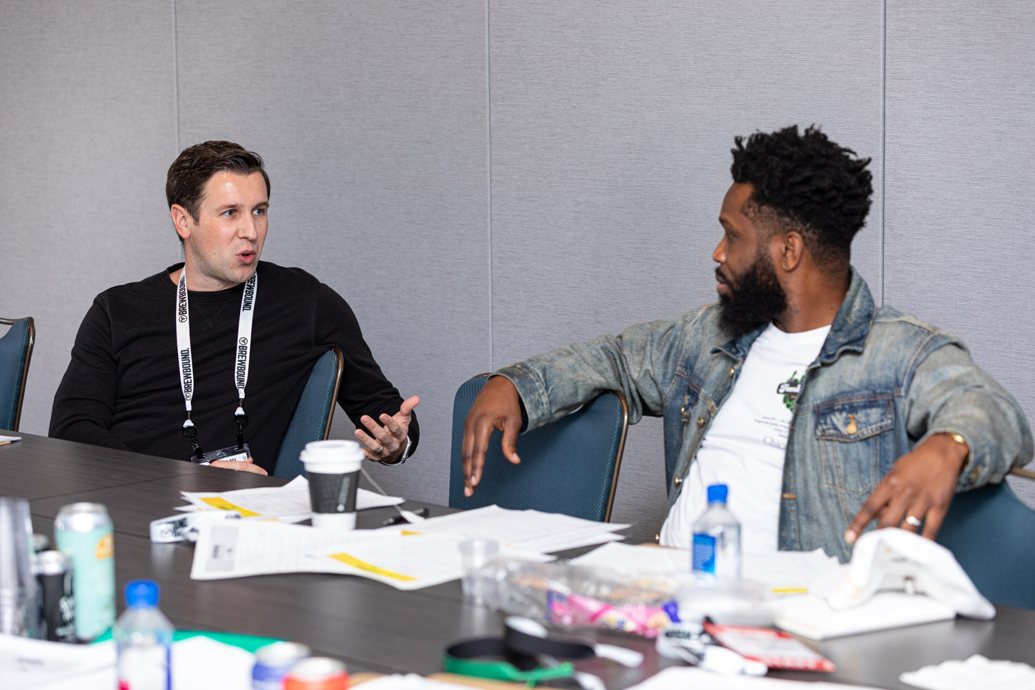 A man in a black sweater with a lanyard and a bearded man in a denim jacket talk at a long gray table with papers and cans.