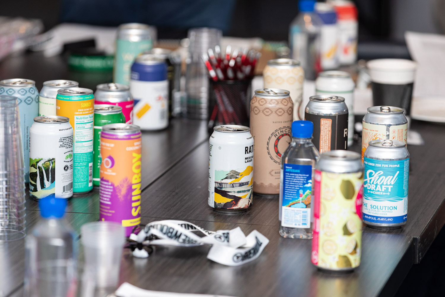 Close view of a table crowded with colorful beverage cans, water bottles and a conference lanyard, with Shoal Draft and Sunboy cans visible.