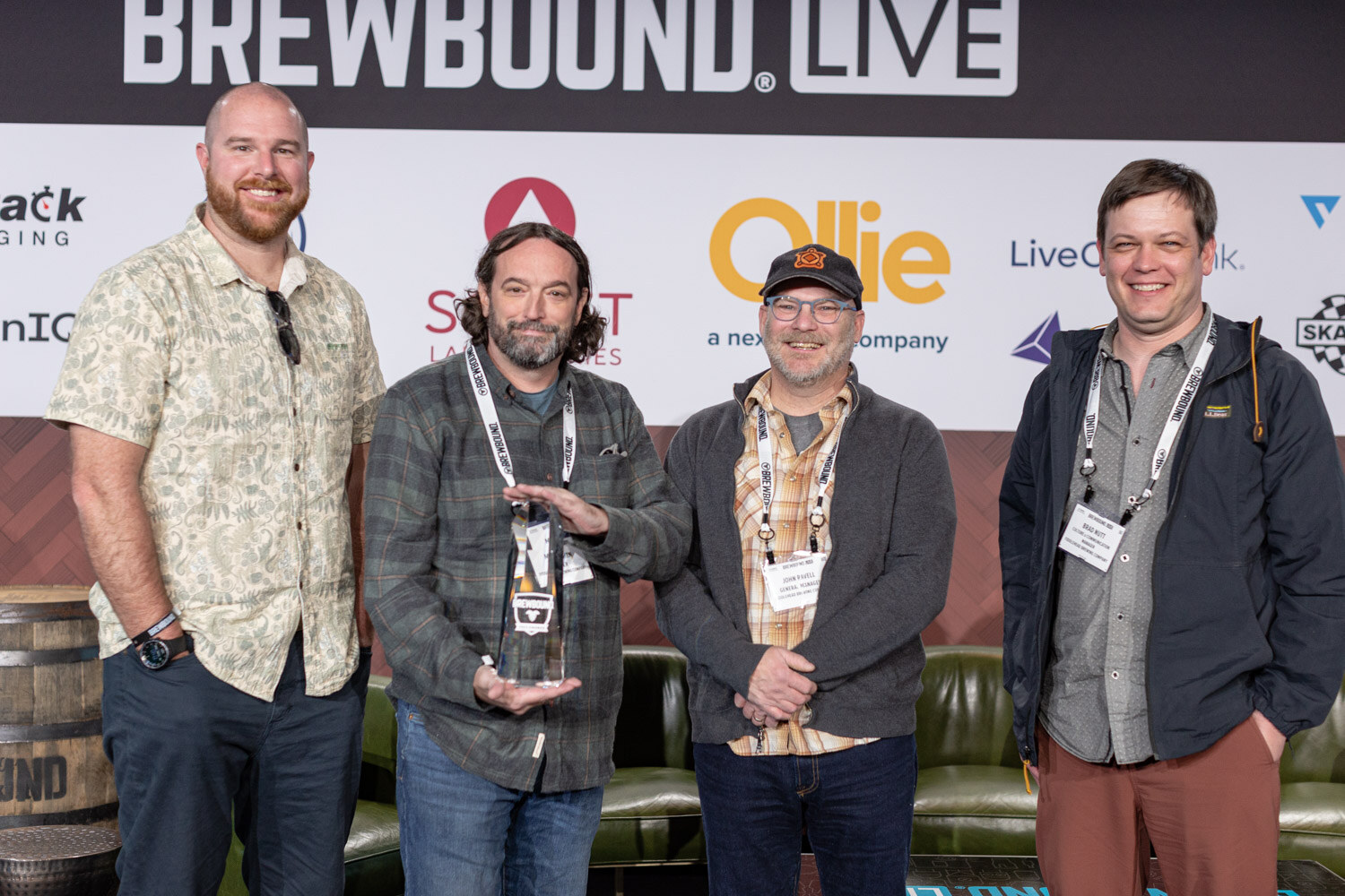 Four men in casual jackets and shirts stand in a row on stage, one holding an award, in front of the Brewbound Live sponsor wall.
