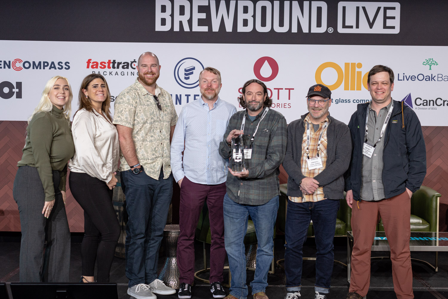 A group of seven people stand in a row on stage in front of the Brewbound Live sponsor wall, one holding an award.
