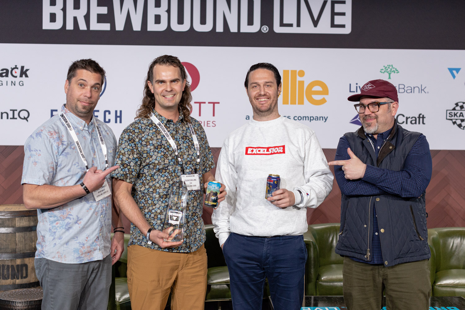 Four men on stage in patterned shirts and jackets, one holding a glass award and another a can, pointing and smiling.