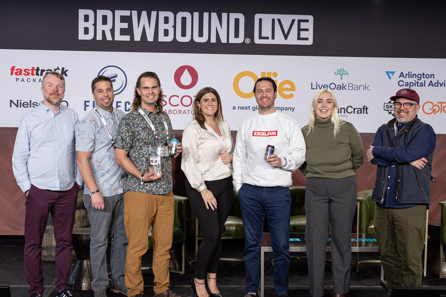 A line of eight people in casual clothes standing in front of the Brewbound Live sponsor wall, several holding trophies and cans.