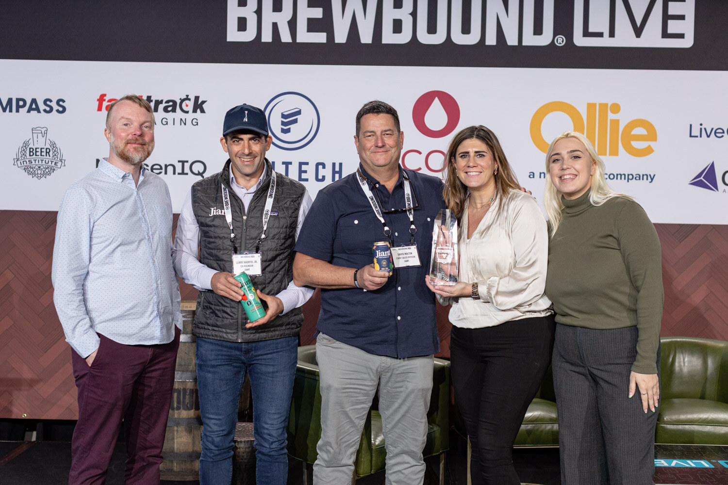 Four people stand together on stage, two men holding cans and a woman holding a glass award, under the Fasttrack and Scott Laboratories logos.
