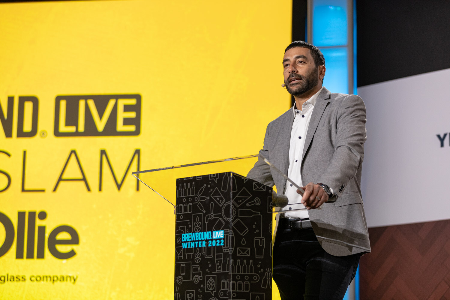 A man in a gray blazer and white shirt speaks at a clear podium in front of a yellow screen reading Slam and Ollie.