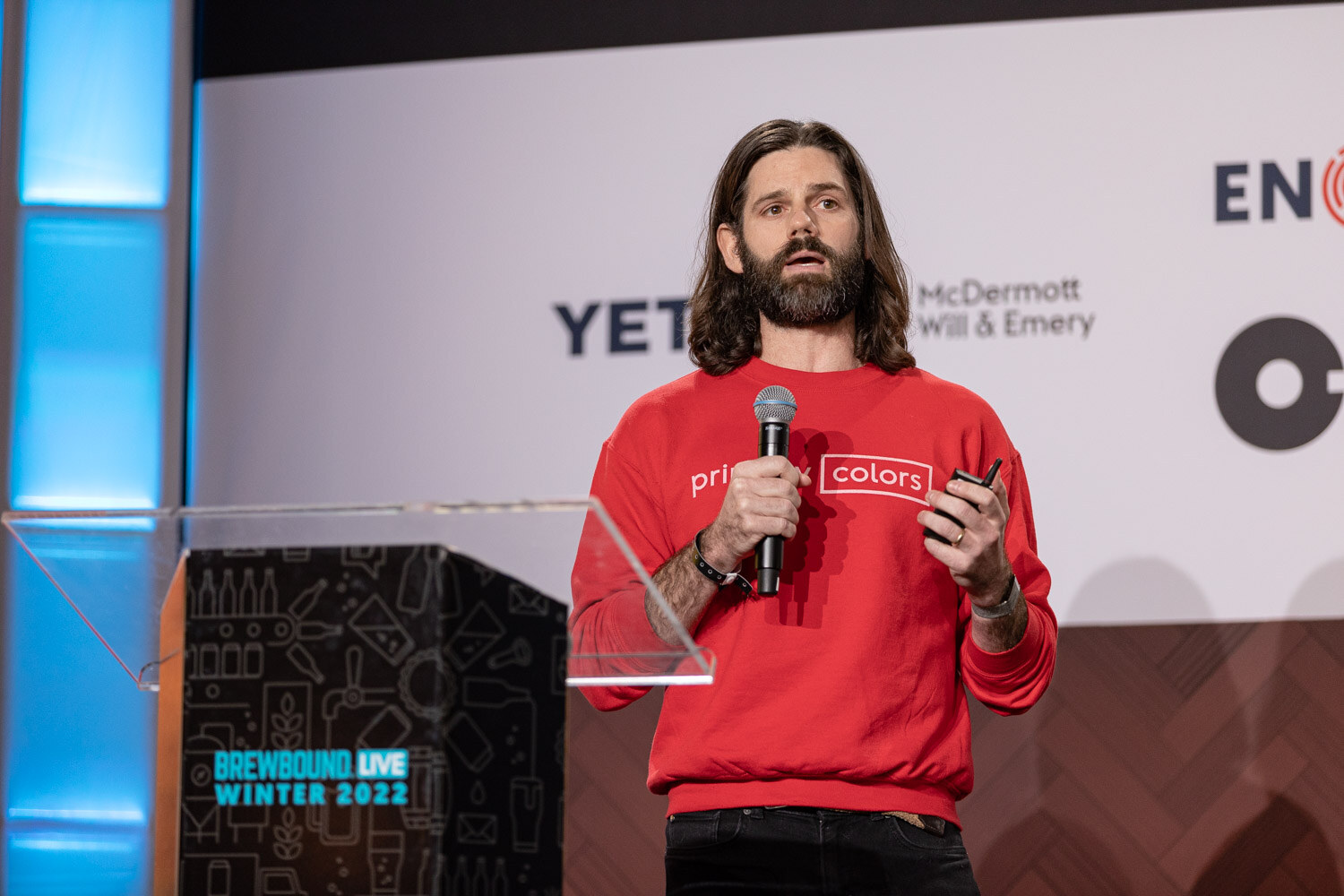 A man in a red sweatshirt reading primary colors holds a microphone and clicker beside a clear podium marked Brewbound Live Winter 2022.