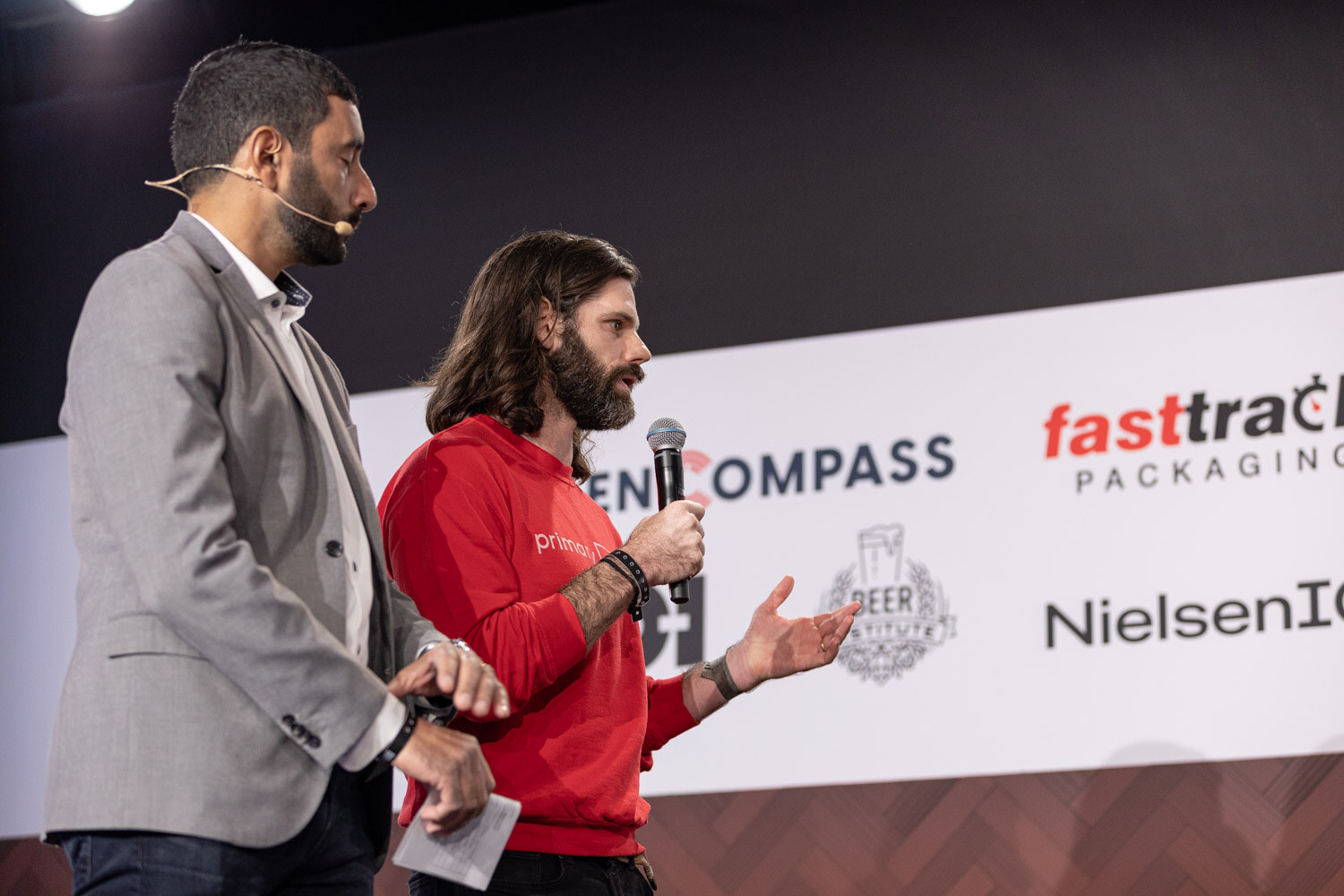 A man in a gray blazer stands next to a long-haired man in a red sweatshirt who gestures with one hand while holding a microphone.