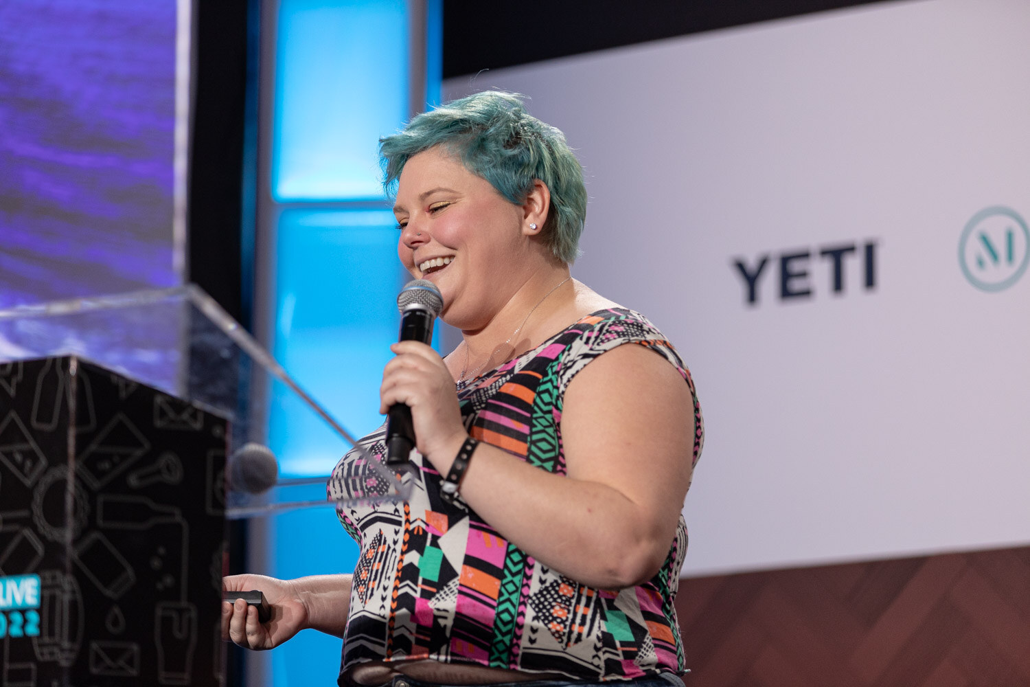 A woman with teal-dyed short hair in a bright patterned top holds a microphone and smiles at the podium.