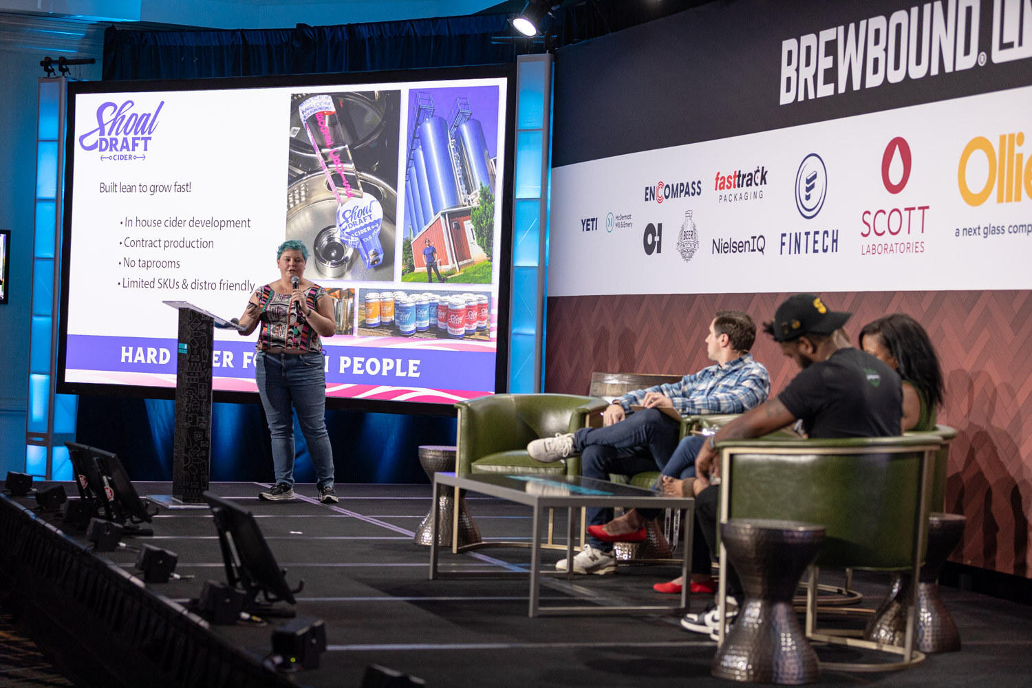 Wide stage view with a presenter at a podium next to a large screen reading Hard Cider for Fun People, with judges seated at right.