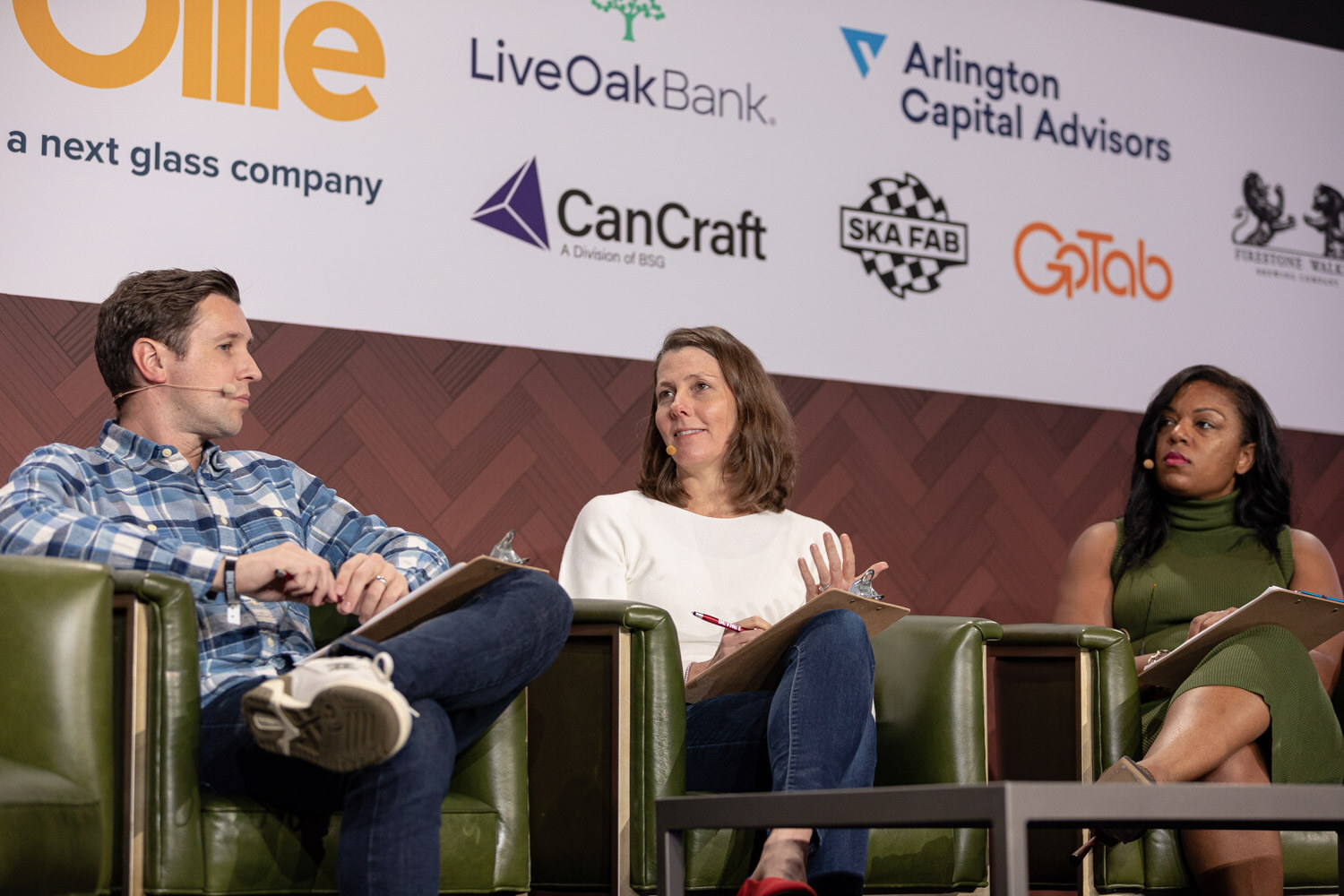 A man in a blue plaid shirt, a woman in a white sweater and a woman in a green sleeveless top sit in armchairs with clipboards in front of sponsor logos.