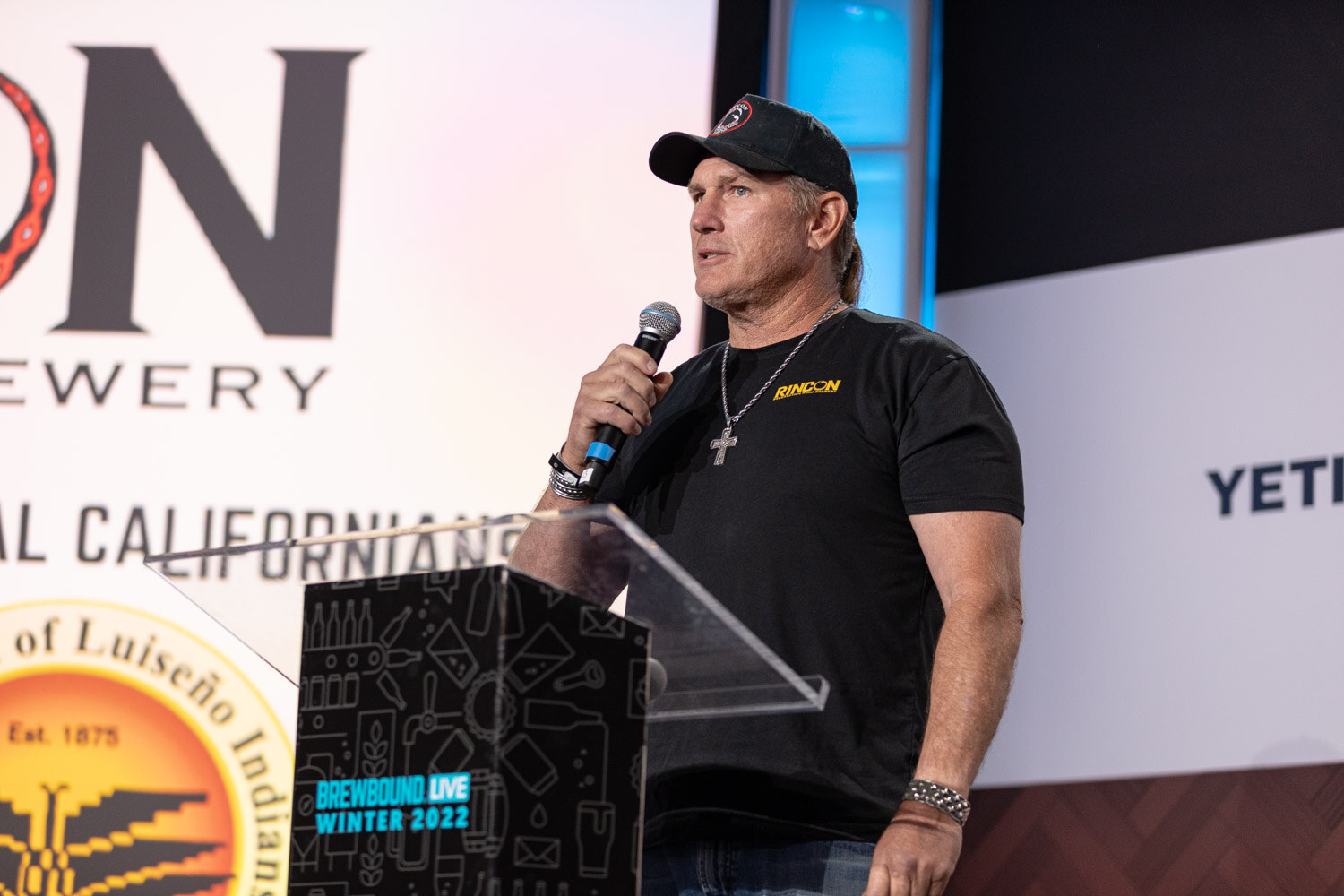 A man in a black cap and black T-shirt speaks into a microphone beside a podium, with a slide showing a Luiseno Indians seal and Rincon logo.