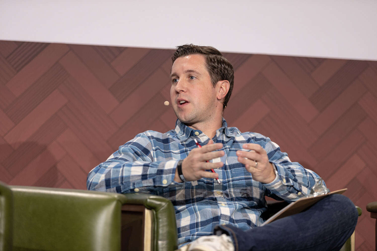 A man in a blue plaid shirt speaks with both hands raised while seated with a clipboard in a green armchair.