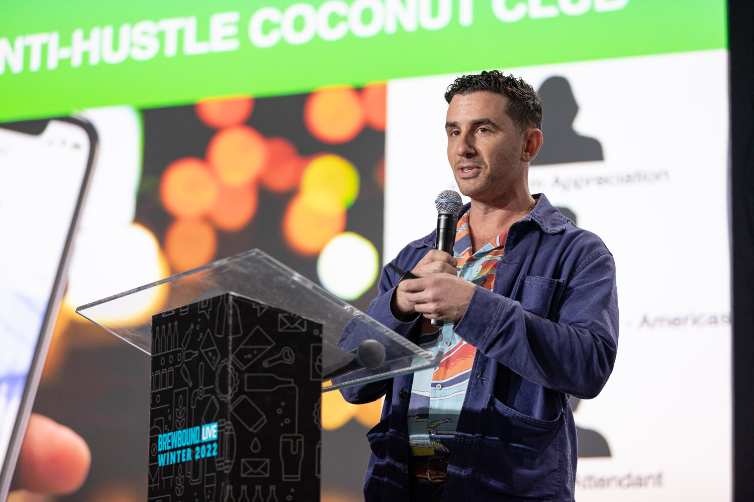 A man in a navy jacket holds a microphone and a clicker next to a large screen with a phone mockup and the words Anti-Hustle Coconut Club.
