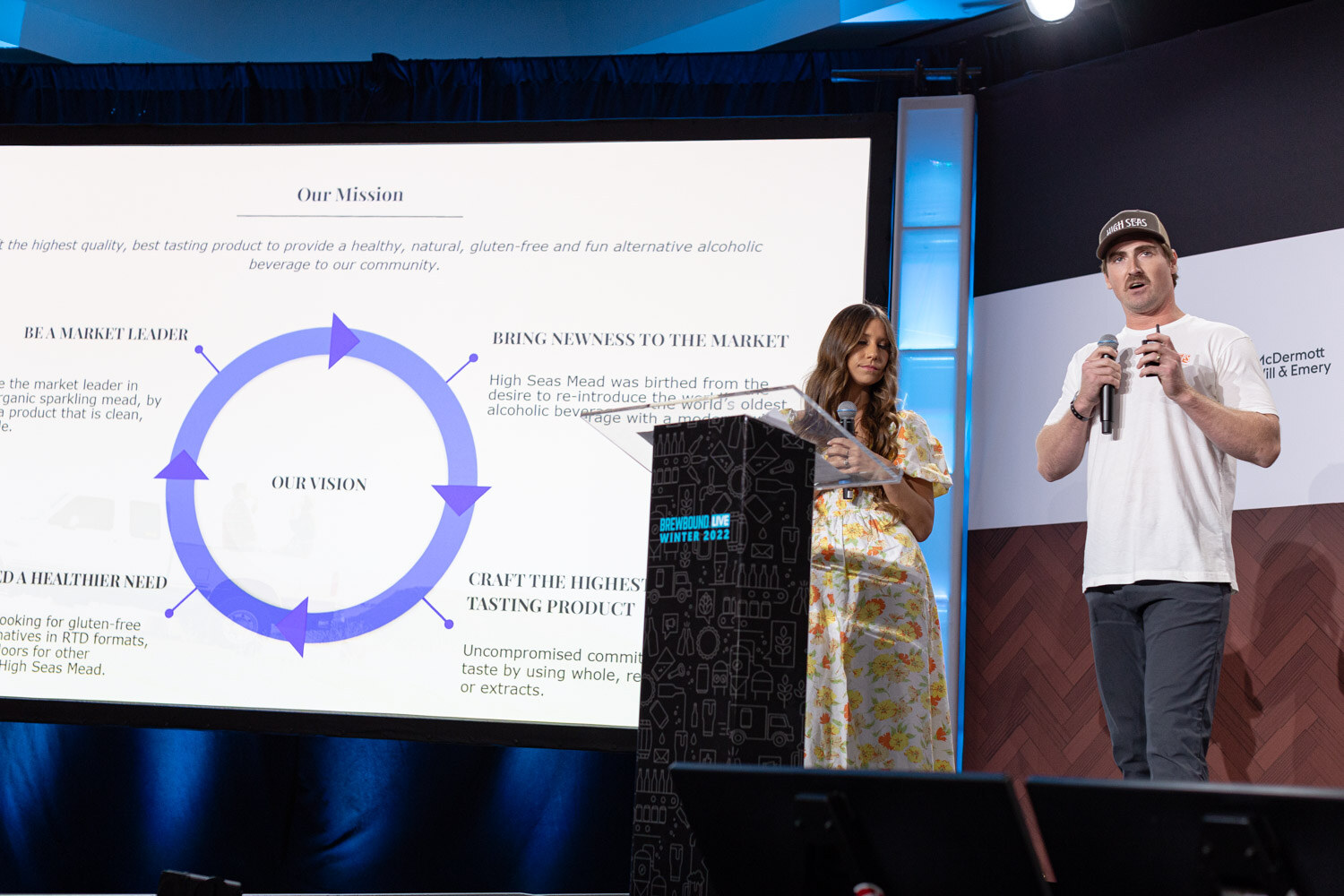 Two presenters stand at the left of a podium next to a large screen showing a circular Our Mission diagram.