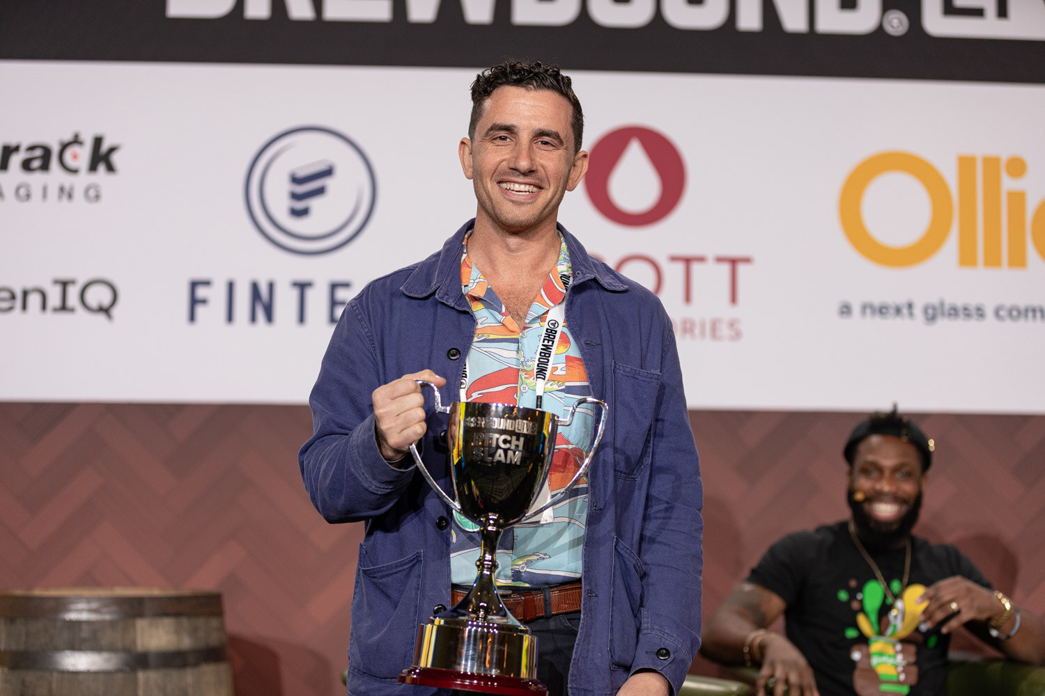A smiling man in a navy jacket over a bright printed shirt holds a large silver trophy cup while a man sits behind him.