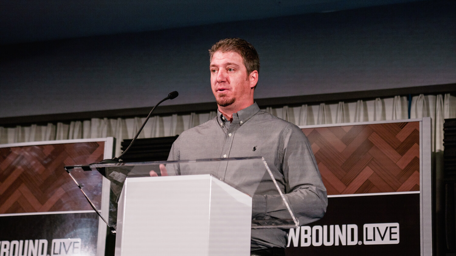 A man in a gray shirt stands behind a clear podium on a small stage with a Brewbound Live sign and herringbone wood panels.