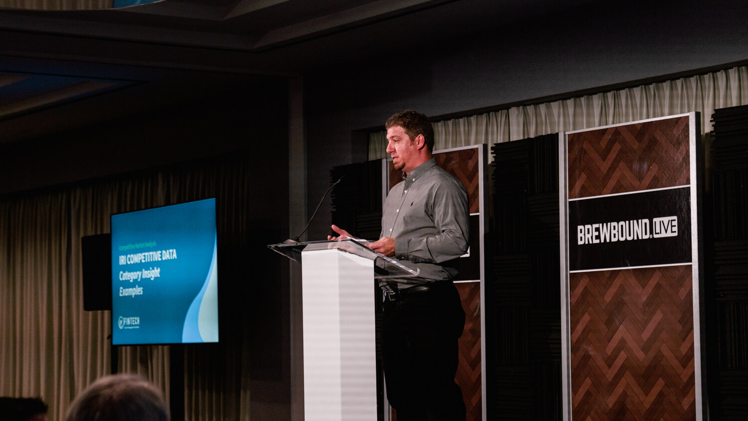 A man at a podium beside a screen displaying a blue slide titled Competitive Data, Category Insight Examples, in a dim ballroom.