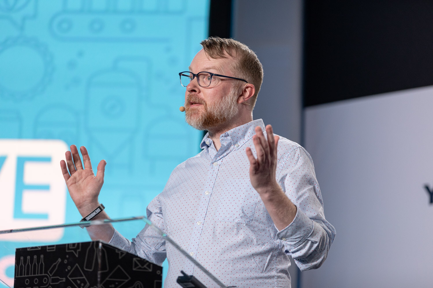 A man with glasses and a beard in a light blue shirt speaks with raised hands at a podium in front of a blue Brewbound Live backdrop.