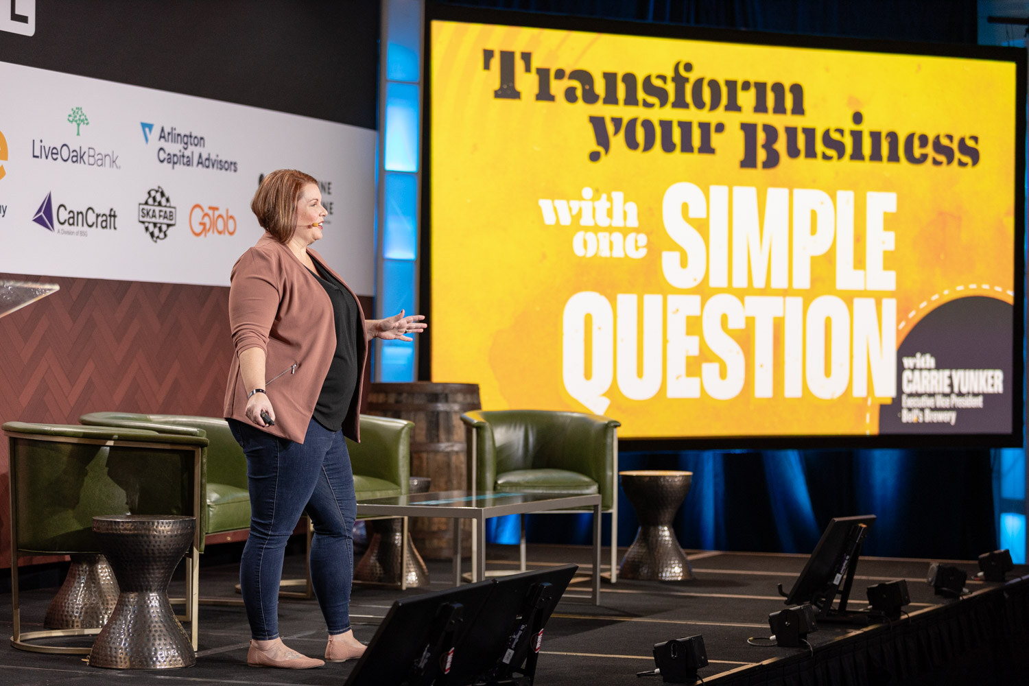 A woman in a pink blazer stands on a large stage beside a yellow screen reading Transform Your Business with One Simple Question.