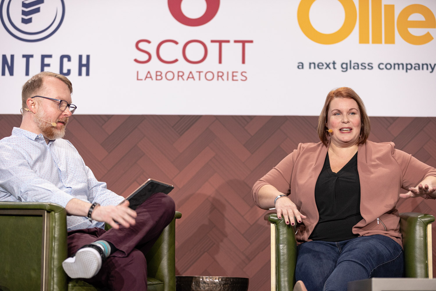 A man with a tablet and a woman in a pink blazer smile at each other in green armchairs in front of the Fintech, Scott Laboratories and Ollie sponsor logos.