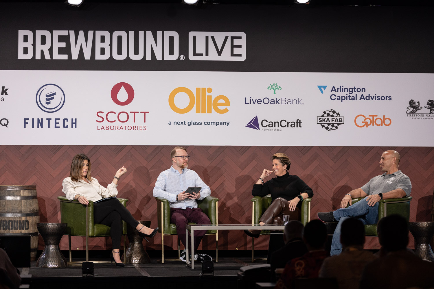 Wide view of four people seated in green armchairs on stage under a Brewbound Live sponsor wall.