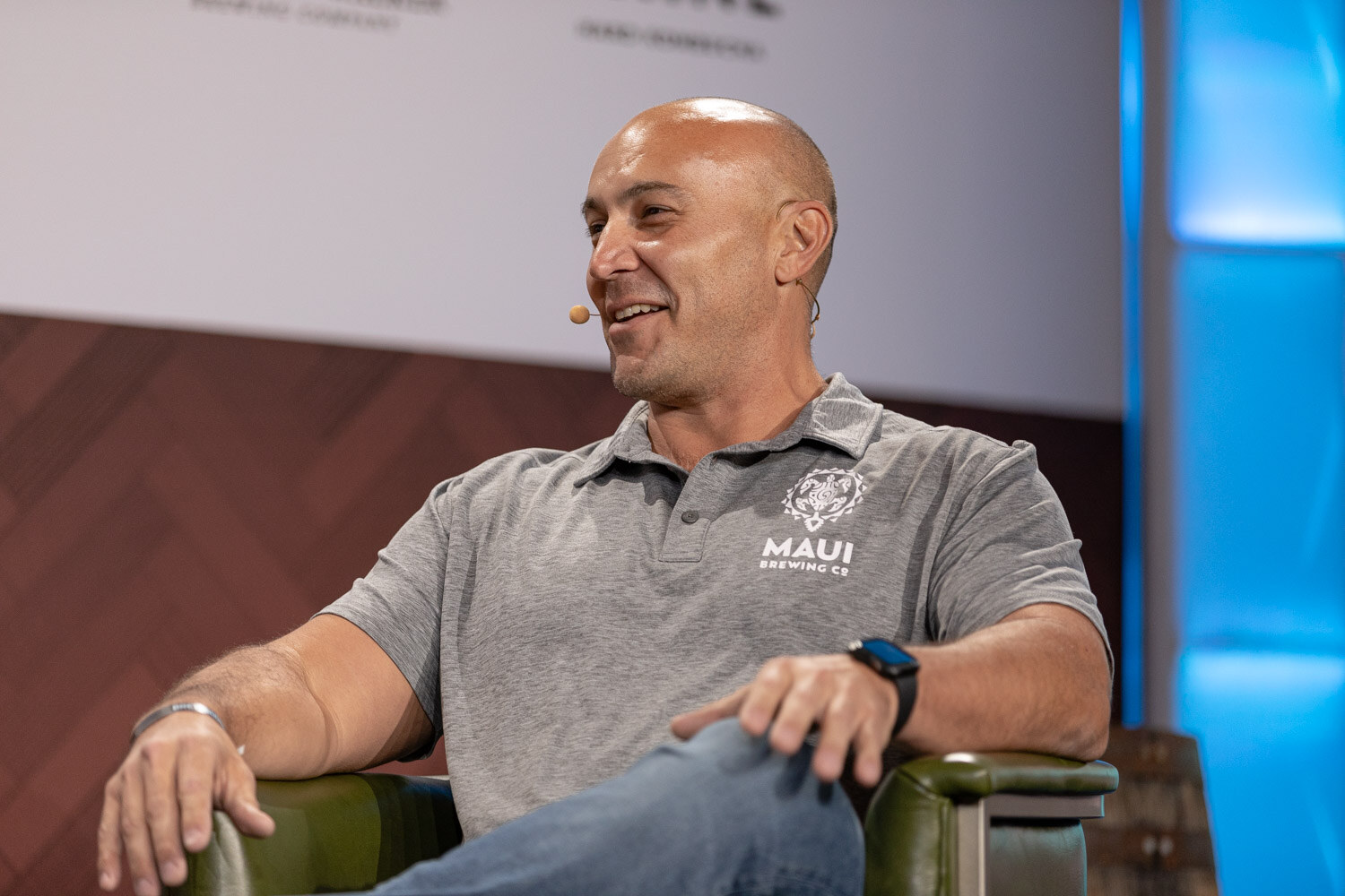 A bald man in a gray polo with a Maui Brewing logo leans back in a green armchair and smiles.