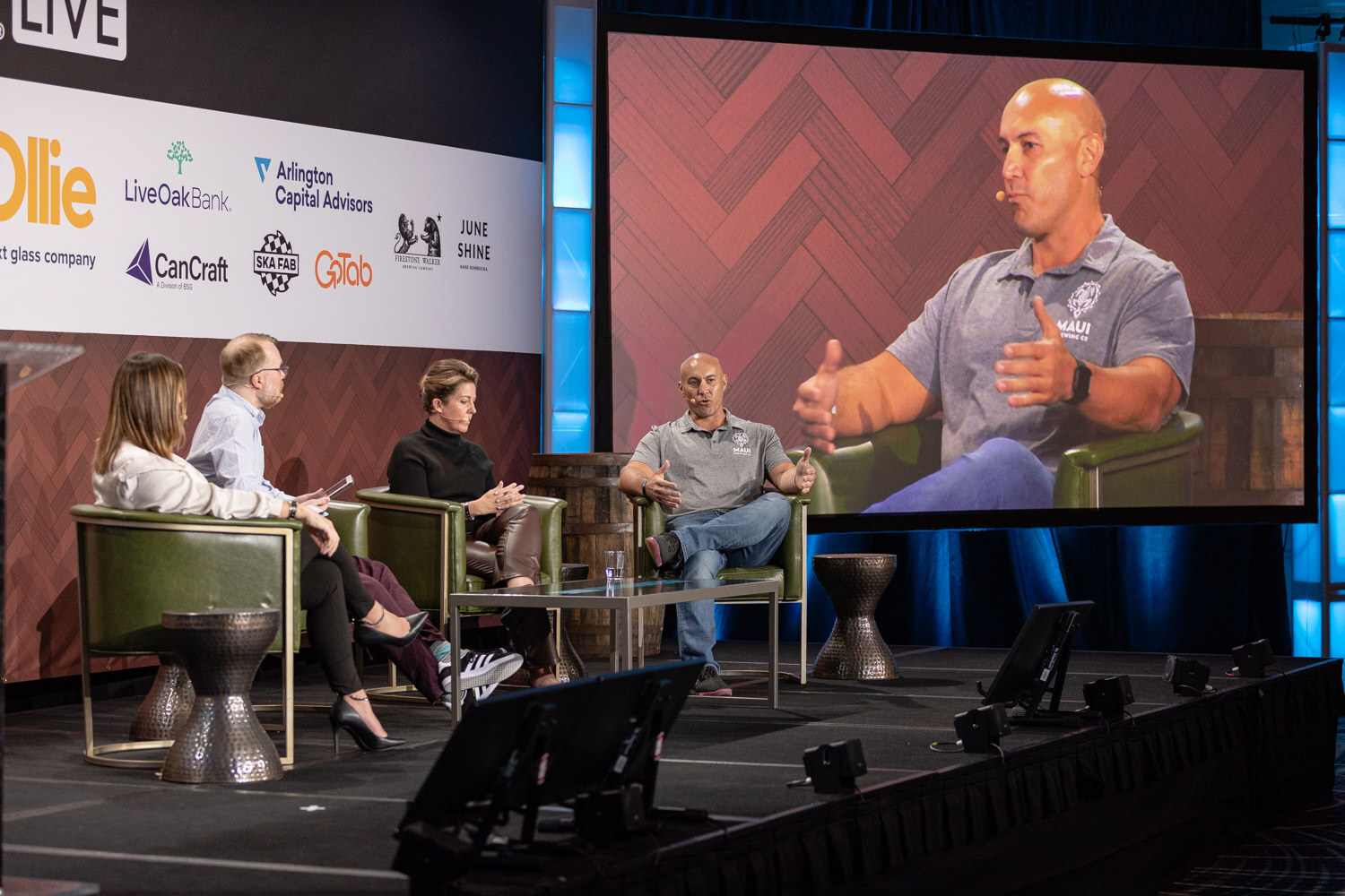 Stage view of four people in armchairs beside a big screen showing a close-up of a bald man in a gray polo with a Maui logo.