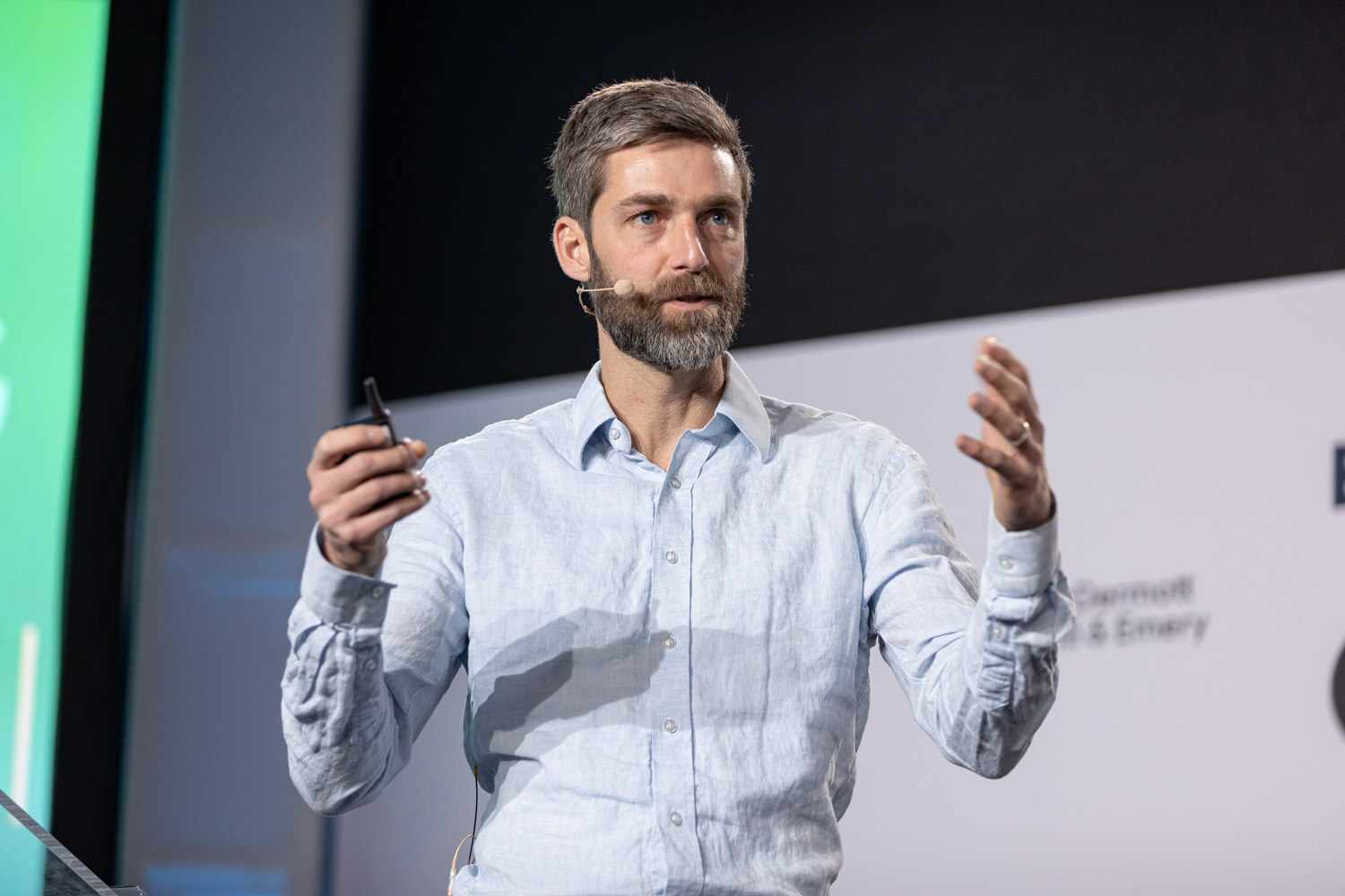 A bearded man in a light blue linen shirt holds a clicker and gestures with both hands on stage.