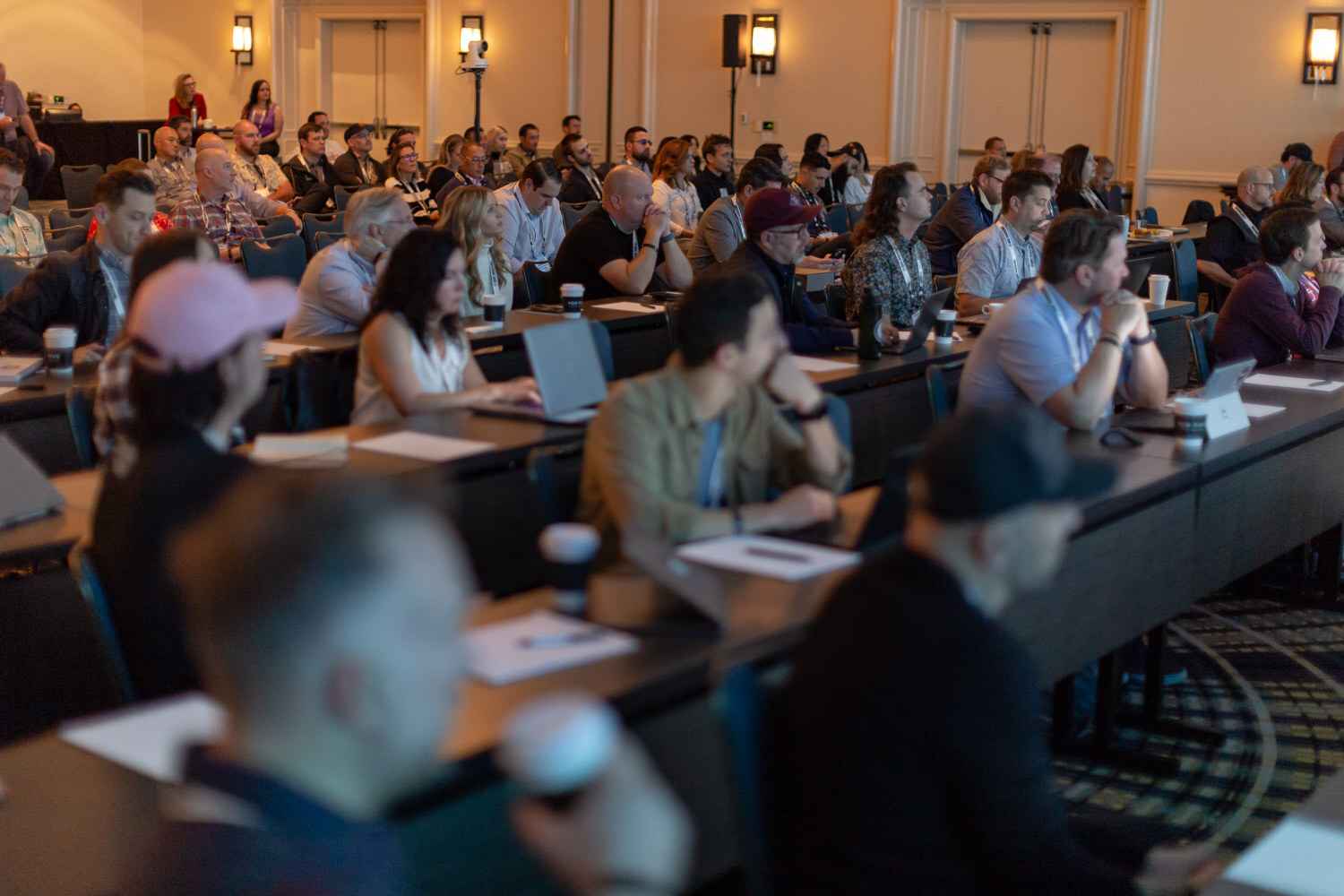 View across rows of attendees seated at classroom-style tables in a warmly lit hotel ballroom.