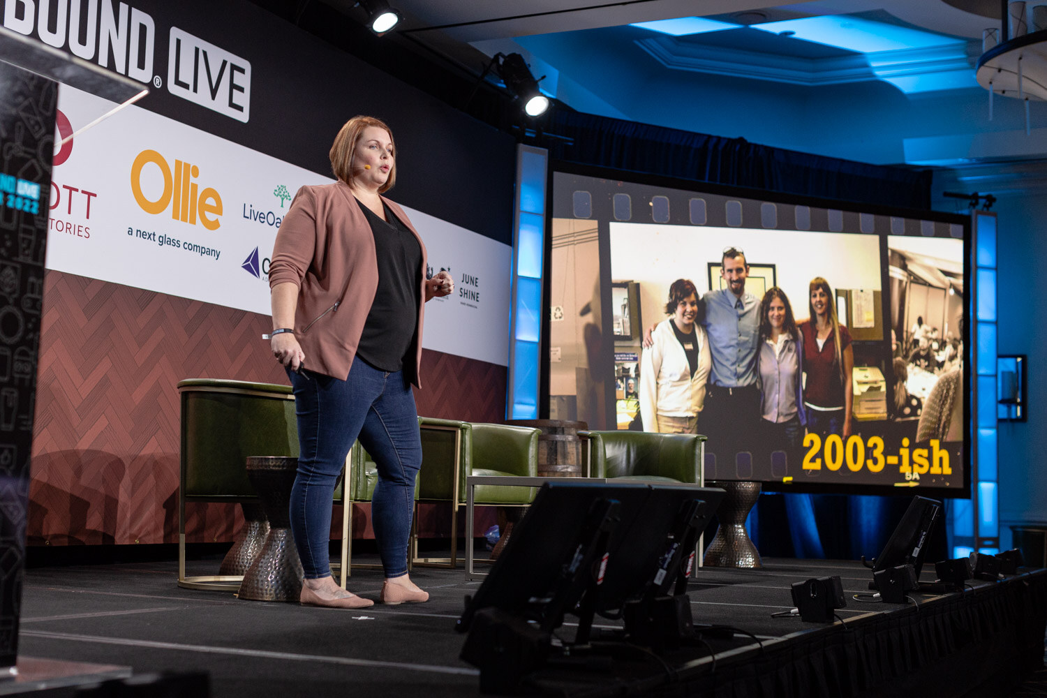A woman on stage beside a large screen showing a group photo of smiling people captioned 2003-ish.