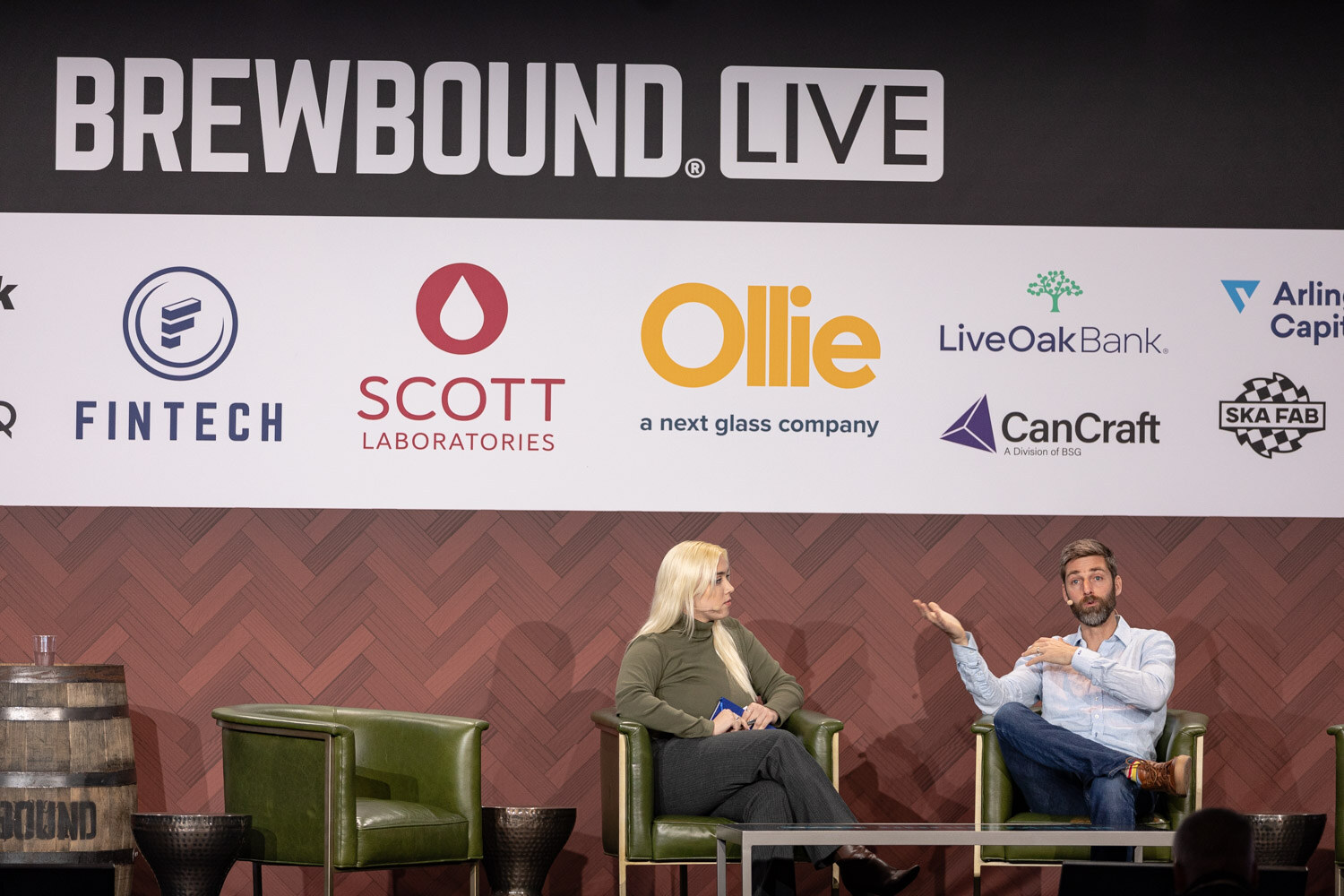 Two people seated in green armchairs on a stage beneath a Brewbound Live banner, the man gesturing with one hand.