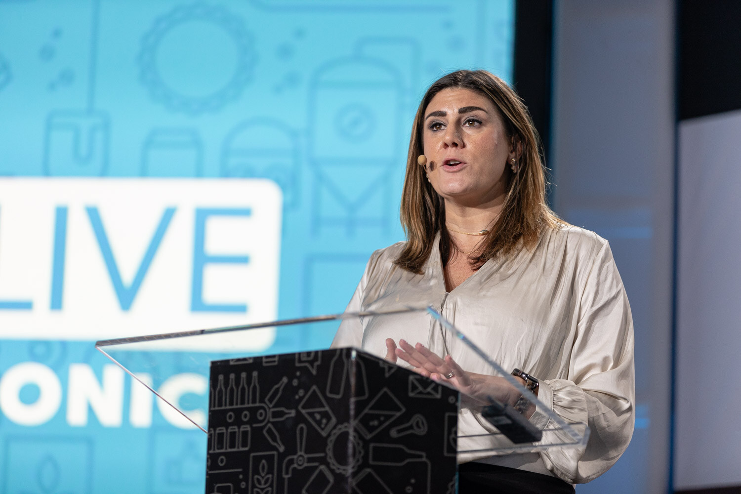 A woman with shoulder-length brown hair in a cream blouse speaks at a clear podium in front of a blue Brewbound Live Santa Monica backdrop.