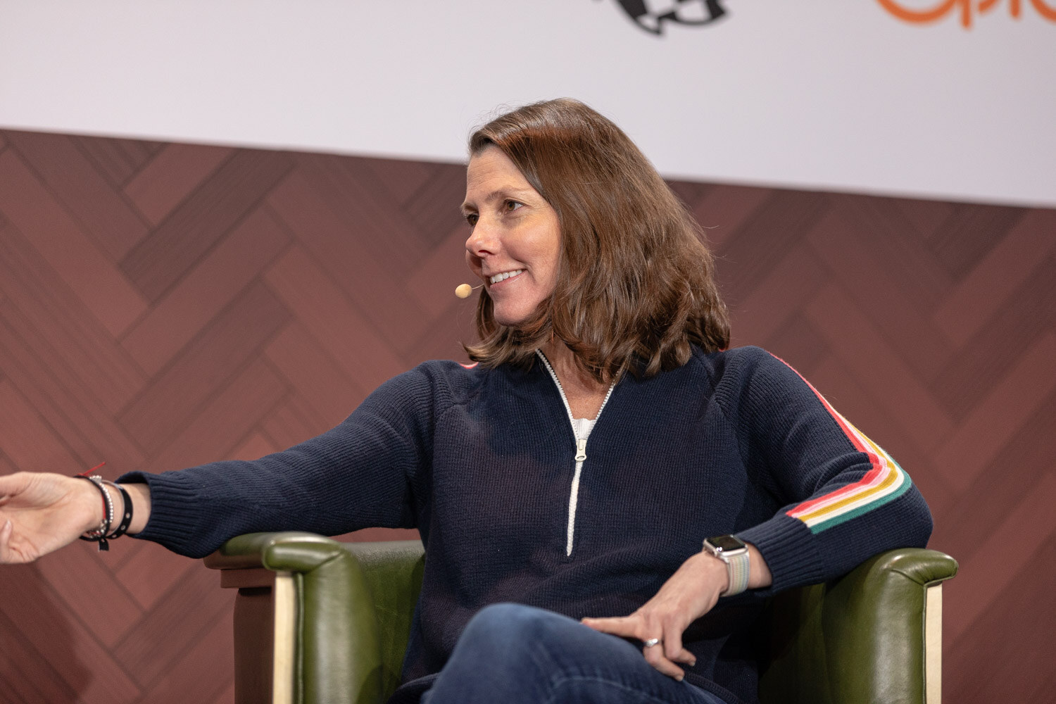 A woman with shoulder-length brown hair in a navy quarter-zip with a rainbow sleeve stripe gestures with an open hand.