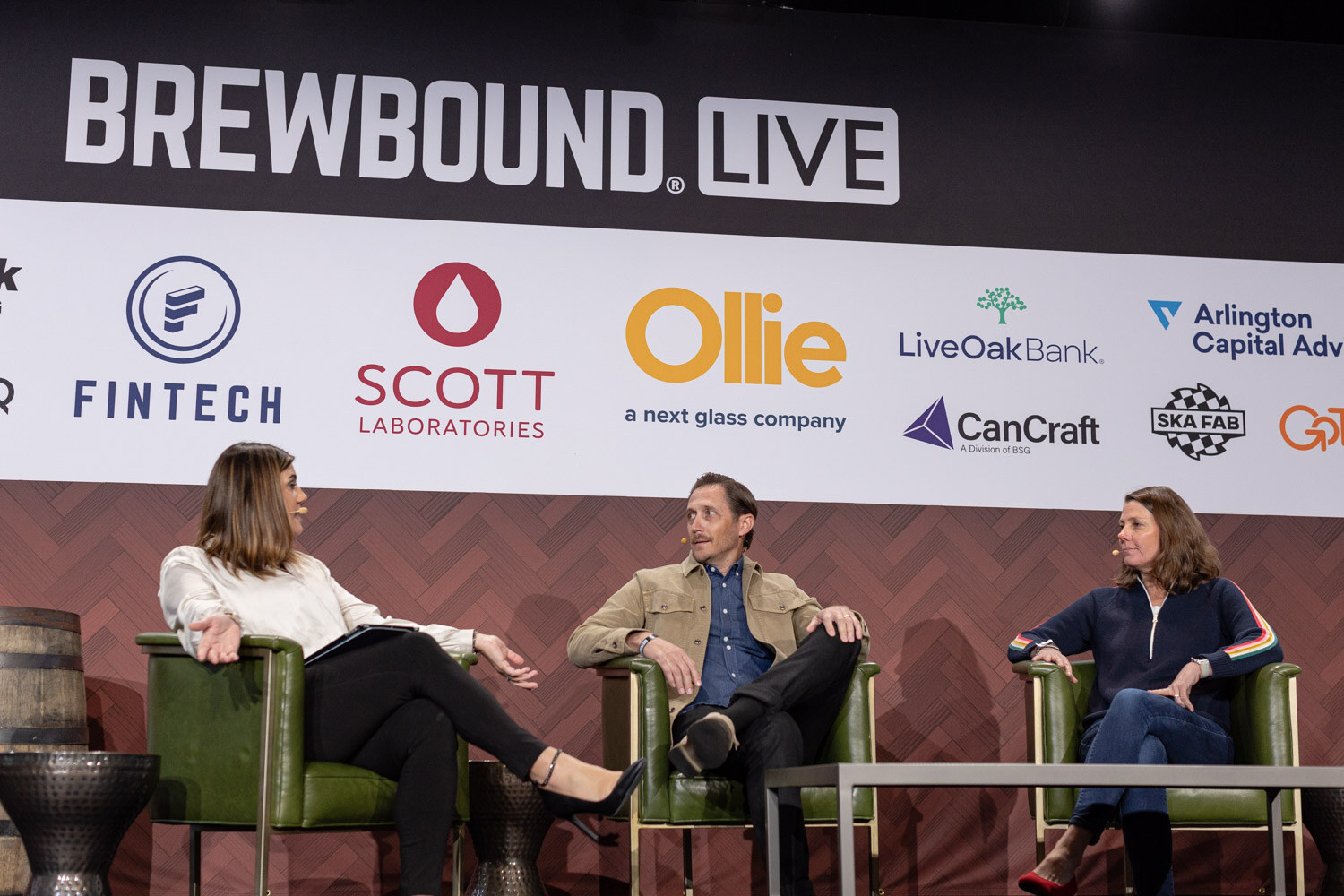 Wide view of a woman in a cream blouse, a man in a tan jacket and a woman in a navy sweater seated on stage under the Brewbound Live sponsor wall.