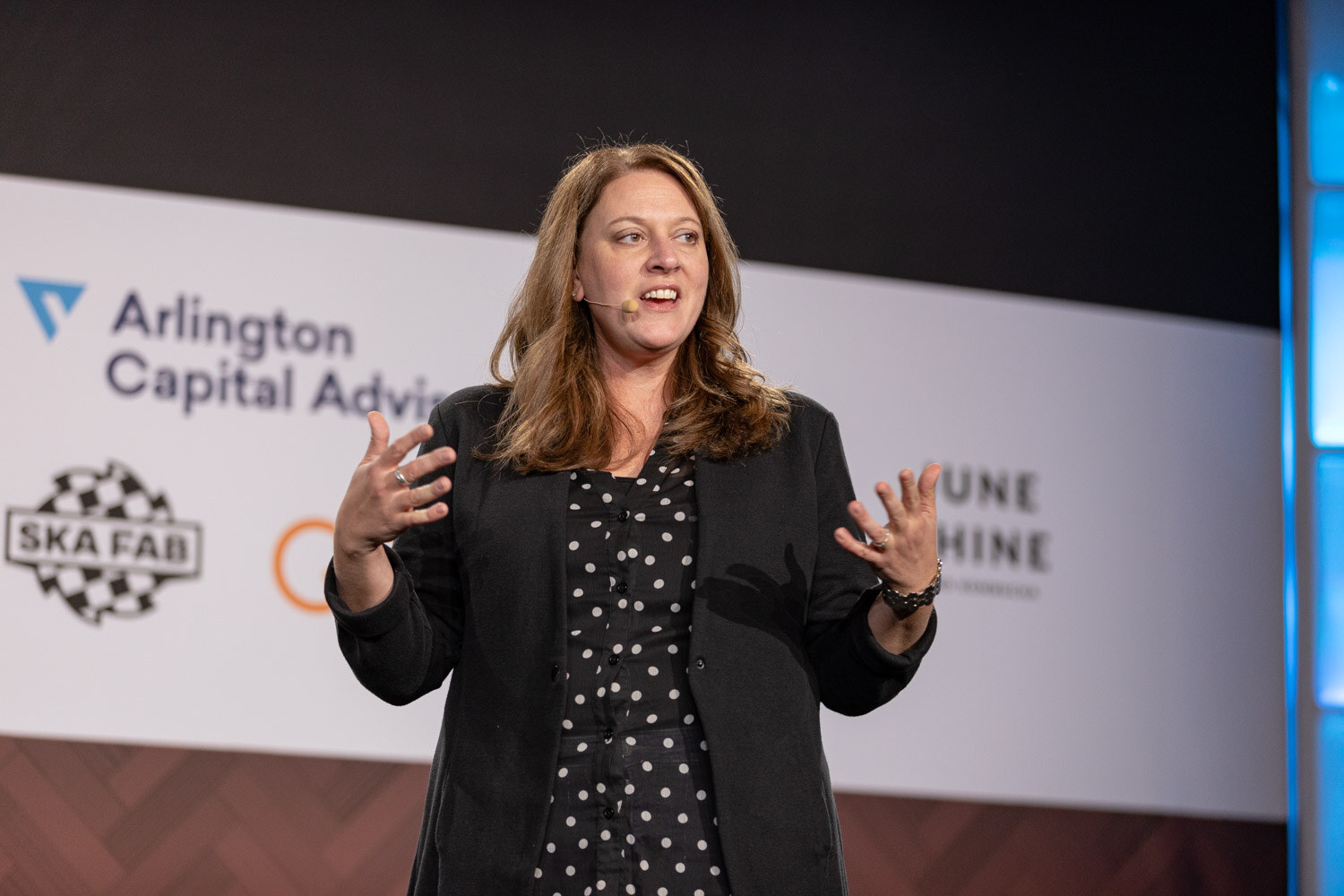 A woman with long brown hair and a headset microphone gestures in front of the Arlington Capital Advisors and Ska Fab sponsor backdrop.