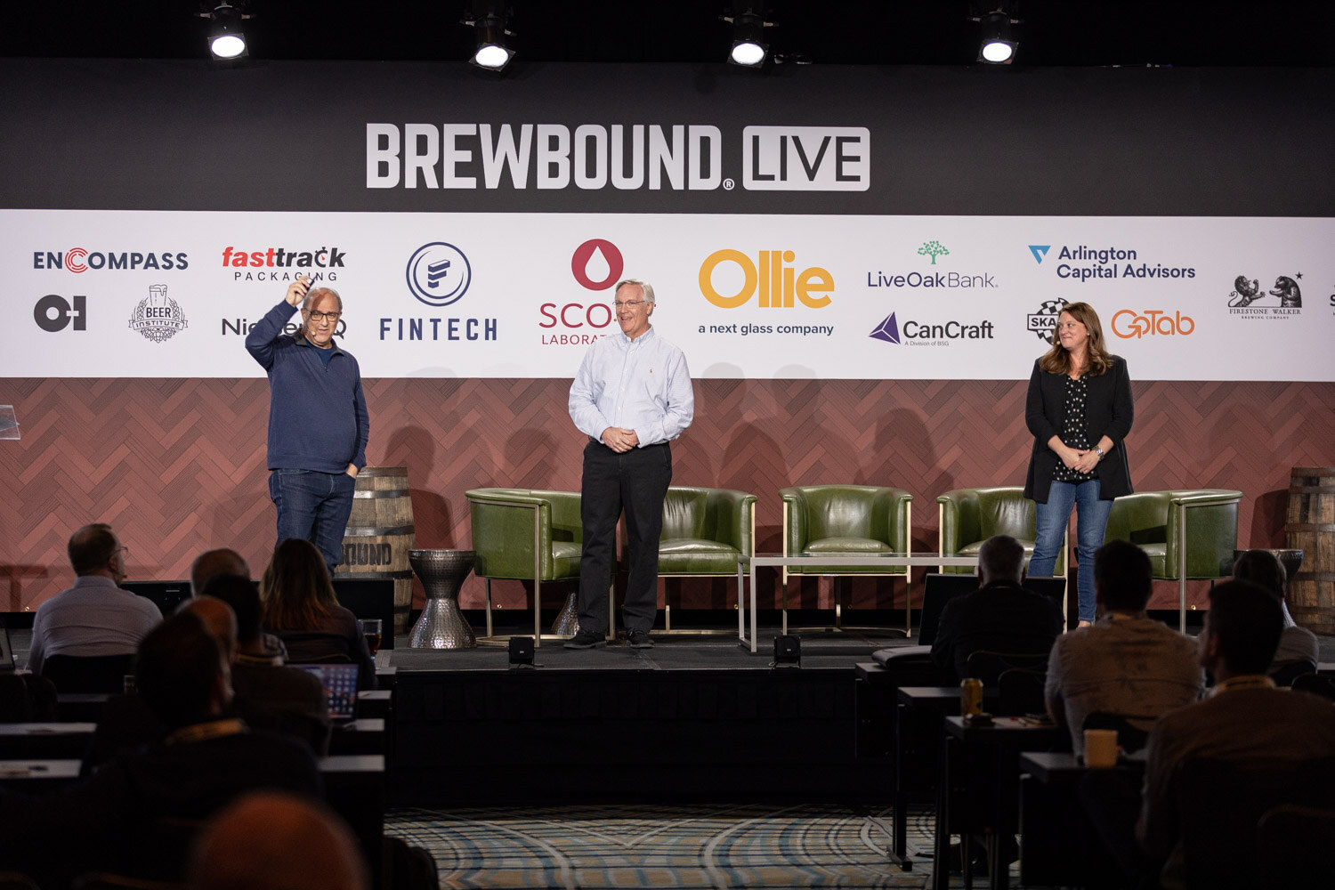 Wide shot of a man in a navy sweater, a man in a white striped shirt and a woman in a black blazer standing on a stage under the Brewbound Live banner.