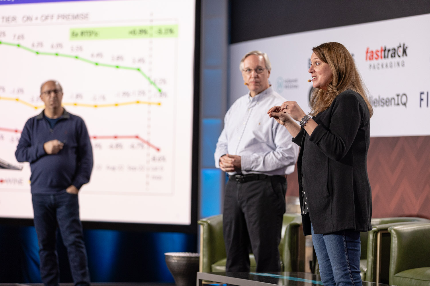 A man in a navy sweater, a man in a white shirt and a woman in a black blazer stand beside a large screen of colorful line charts.