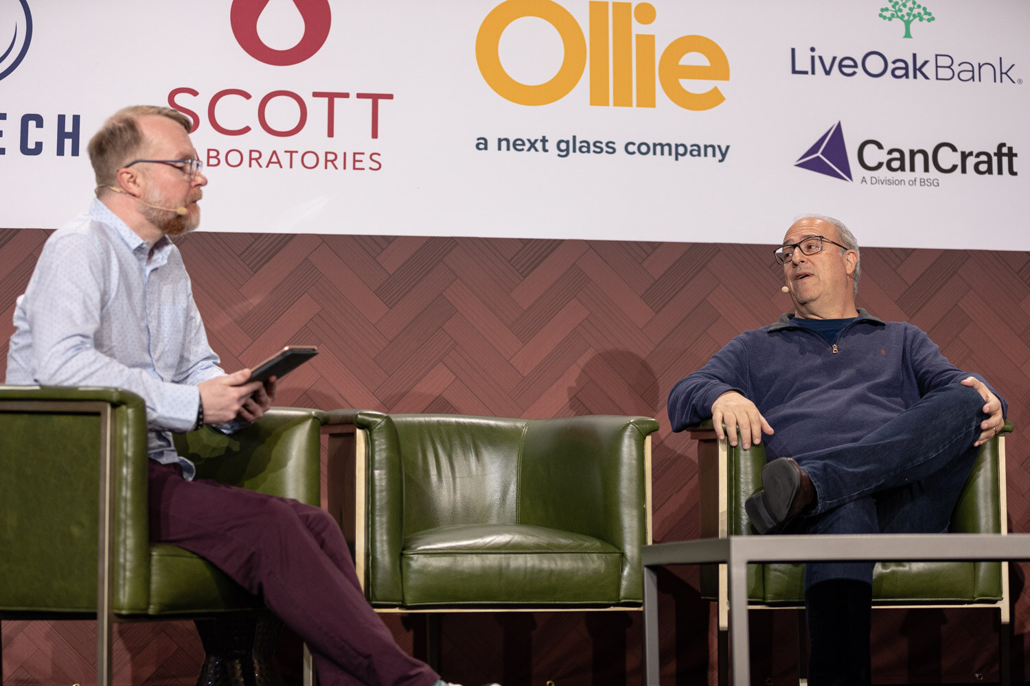 A man with a tablet and light blue shirt sits next to a man in a navy quarter-zip with sponsor logos including Scott Laboratories and CanCraft behind them.